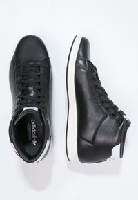 Svarta läder hög-top sneakers med vit sula, som har svarta snörningar, perforerade sidor och en märkt tunga. Det finns även en liten röd etikett som accent.