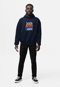 Felpa navy con grafica Pepsi sulla parte frontale. Abbinata a pantaloni a quadri neri e scarpe scure con dettagli bianchi.