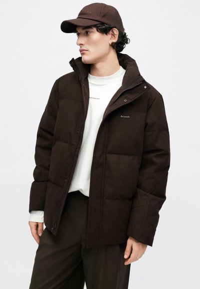 Brūna pūdera jaka ar augstu apkakli, rāvējslēdzēju priekšpusē un sprādzēm. Valkāta ar baltu kreklu un brūnu cepuri ar logotipu.