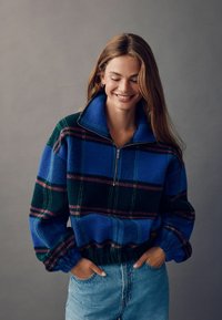 Blauer und grüner karierter Fleece-Pullover mit einem halbseitigen Reißverschluss, lockerem Schnitt, elastischen Bündchen und einem gerafften Saum. Das Model trägt hellblaue Jeans.