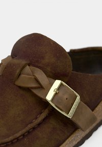 Καφέ σουέντ παπούτσι τύπου slip-on με πλεγμένο δερμάτινο λουράκι και λεπτομέρεια με χρυσή αγκράφα, διαθέτοντας υφή στην επιφάνεια και σχεδιασμό με στρογγυλεμένη μύτη.