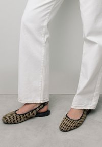 Des chaussures slingback noires et beiges à motif avec une semelle plate, dotées d'un bout arrondi et d'un talon élastiqué, portées avec un pantalon blanc à jambe droite.