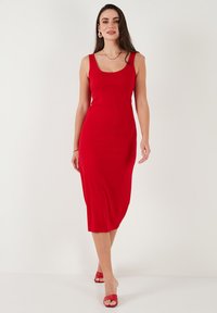 Abito midi rosso aderente con scollatura a barca e spalline larghe. Texture liscia, forma bodycon, abbinato a tacchi aperti rossi coordinati.