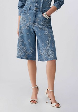 Jeans bermuda kratke hlače z belo paisley vezenino, kombinirane z ujemajočo se jeans srajco in belimi sandali z visokimi petami.