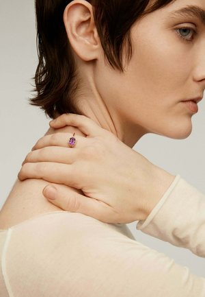 Primer plano de una persona con cabello corto castaño tocándose el cuello, vistiendo una blusa beige y un anillo dorado con una gran piedra preciosa púrpura en el dedo.