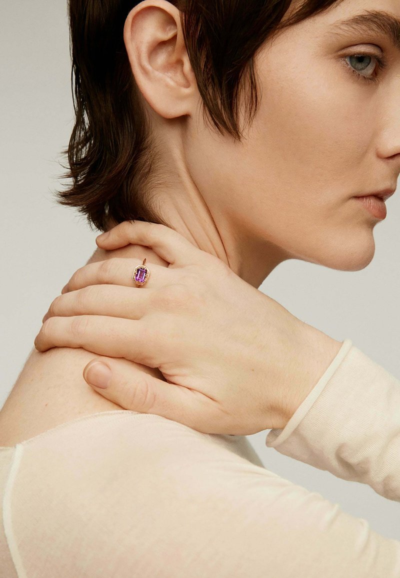 Primer plano de una persona con cabello corto castaño tocándose el cuello, vistiendo una blusa beige y un anillo dorado con una gran piedra preciosa púrpura en el dedo.