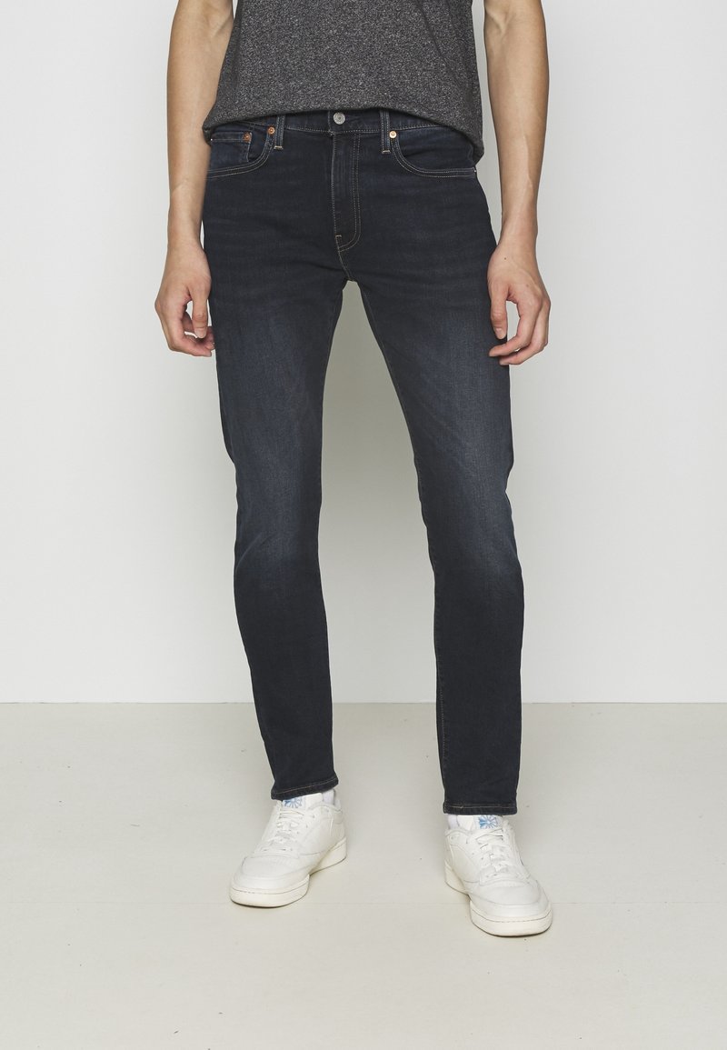 Personne portant un jean slim bleu foncé, un t-shirt gris et des baskets blanches, debout devant un fond clair uni.