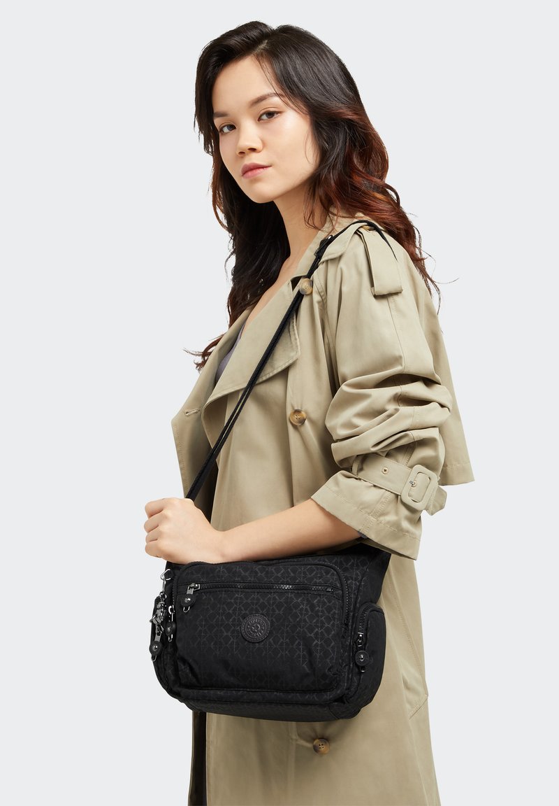 Schwarze Schultertasche aus strukturiertem Stoff, mit Reißverschlussfächern und Logodetail. Quer über die Schulter getragen mit verstellbarem Gurt vor einem hellen Hintergrund.