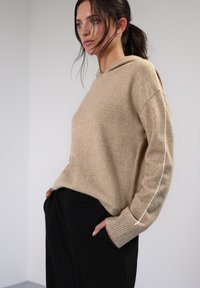 Beige gestrickter Kapuzenpullover mit gerippten Bündchen und kontrastierenden weißen Streifen an den Ärmeln. Kombiniert mit schwarzen Hosen, die eine lockere Silhouette präsentieren.