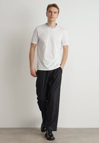 Fehér pamut póló, kerek nyakkal, fekete csíkozott, bő szárú nadrággal párosítva. A modellen fekete cipők vannak. Egyszerű, modern öltözet.