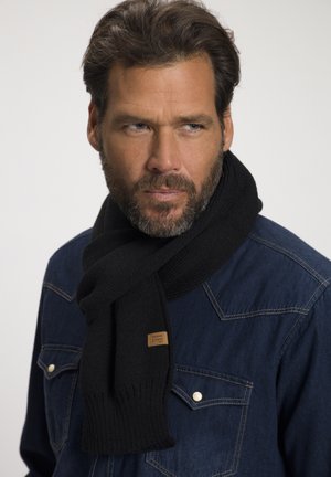 Black knitted scarf with ribbed ends, featuring a small leather tag. Worn over a denim shirt with light-colored stitching.