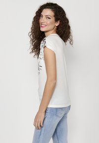 T-shirt blanc à manches courtes avec un design graphique noir sur l'épaule. Tissu doux, coupe décontractée, encolure ronde. Porté avec un jean bleu clair.