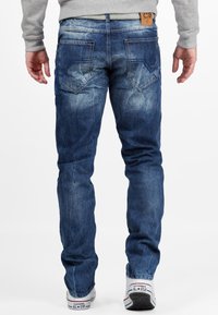 Jean en denim bleu foncé avec un aspect délavé, comportant des poches arrière et une patch en cuir brun au niveau de la taille, associé à des baskets blanches.