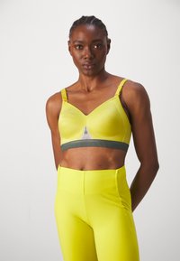 Geel sportbeha met een groene band, voorzien van verstelbare bandjes en een V-vormige halslijn. De stof is glad en rekbaar, met minimale details.