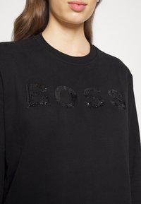 Svart sweatshirt med texten "BOSS" i glänsande svarta paljetter. Rundhalsad design med slät tygtextur och avslappnad passform.