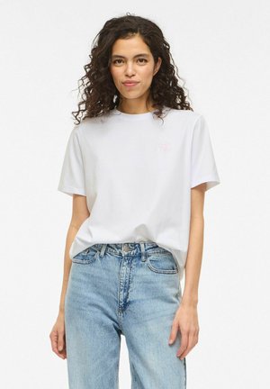 Donna con capelli ricci che indossa una t-shirt bianca con un piccolo disegno di rosa rosa, infilata in jeans a vita alta di colore azzurro chiaro.