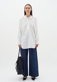 Biela oversized košeľa s vysokým golierom a nariasenými ramenami v kombinácii s voľnými tmavomodrými džínsami. Model drží čiernu tašku.