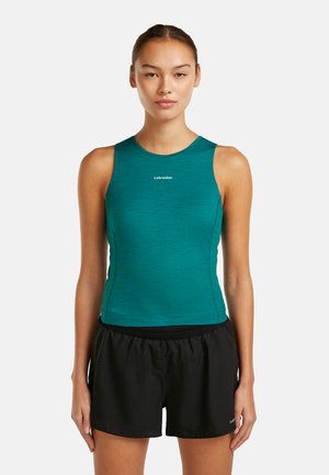 Ung kvinde iført teal ærmeløs træningstop og sorte shorts, stående med front mod kameraet mod en ensfarvet hvid baggrund.