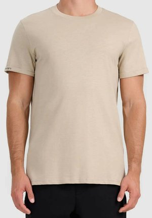 Man die een effen beige korte mouw t-shirt en zwarte broek draagt, staat met ontspannen armen langs het lichaam.