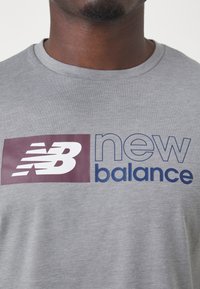 Szara bawełniana koszulka z okrągłym dekoltem. Z przodu znajduje się bordowy kwadratowy logo z białym napisem "NB" oraz niebieskim tekstem "new balance".