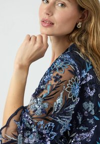 Blusa azul marino transparente con bordado floral intrincado en tonos azules y lentejuelas. Presenta mangas fluidas y detalles delicados en la tela.