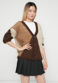 Frau trägt eine übergroße Cardigan in Braun, Beige und Creme mit großen Knöpfen über einem weißen Oberteil und einem schwarzen Plisseerock vor weißem Hintergrund.