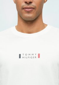 Λευκό βαμβακερό μπλουζάκι με λογότυπο "TOMMY HILFIGER" σε μαύρο και κόκκινο, με στρογγυλή λαιμόκοψη και κοντά μανίκια. Λείο υλικό.
