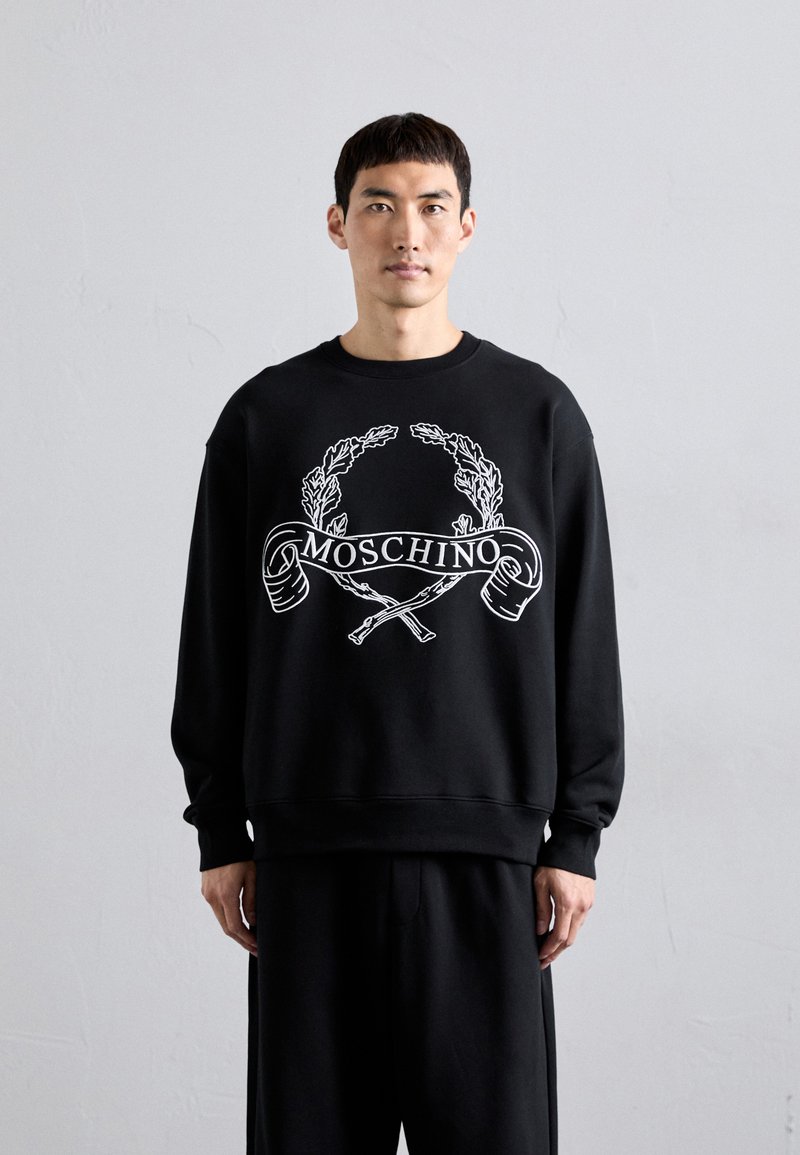 Mann in schwarzem Moschino-Sweatshirt mit weißem Lorbeerkranz- und gekreuztem Zweige-Logo, steht vor einem schlichten hellgrauen Hintergrund.