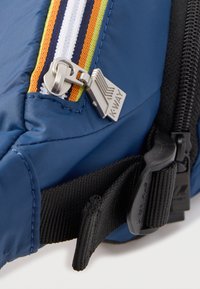 Μπλε υφασμάτινη τσάντα με μεταλλικό φερμουάρ που φέρει την ετικέτα "K-WAY", με μαύρο ρυθμιζόμενο ιμάντα και πολύχρωμη ριγέ λεπτομέρεια στο φερμουάρ.