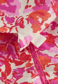 Látka s abstraktnou kvetinovou potlačou v červenej, ružovej a fialovej farbe, s bielou visačkou a detailom zipsu lemovaného ružovou čipkou.