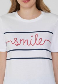 Camiseta de algodón blanca con rayas horizontales azul marino y texto bordado en rojo que dice "smile". Cuello redondo estándar y mangas cortas.