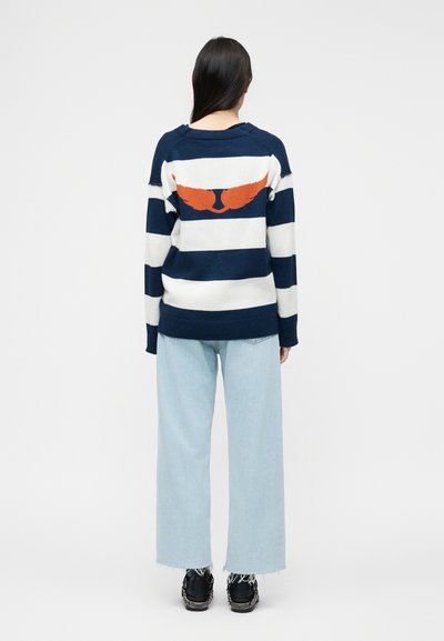Γυναίκα που φορά πουλόβερ με μπλε ναυτικό και λευκές ρίγες, με σχέδιο πορτοκαλί γάτας στην πλάτη, ανοιχτόχρωμα τζιν και μαύρα αθλητικά παπούτσια, στέκεται με την πλάτη προς το μέρος μας.