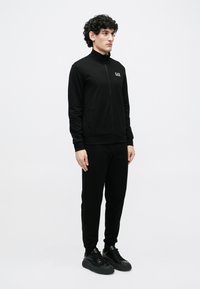 Młody mężczyzna stojący w czarnej kurtce EA7 zapinanej na zamek, dopasowanych czarnych spodniach dresowych i czarnych trampkach na jednolitym białym tle.