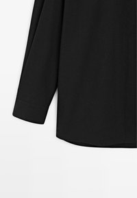 Schwarzes Langarmshirt mit glatter Textur, geschwungenem Saum und geknöpften Manschetten. Der Stoff wirkt leicht und atmungsaktiv, ideal zum Übereinandertragen.