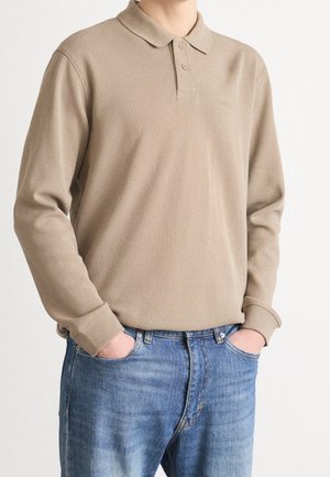 Beżowa koszulka polo z długim rękawem, kołnierzykiem i guzikami, noszona z niebieskimi dżinsami, ręce wsunięte w przednie kieszenie.