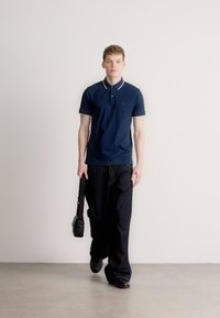 Tricou polo bleumarin cu margine albă la guler, asortat cu blugi largi din denim închis. Modelul ține o geantă mică neagră, stând în fața unui fundal deschis la culoare.