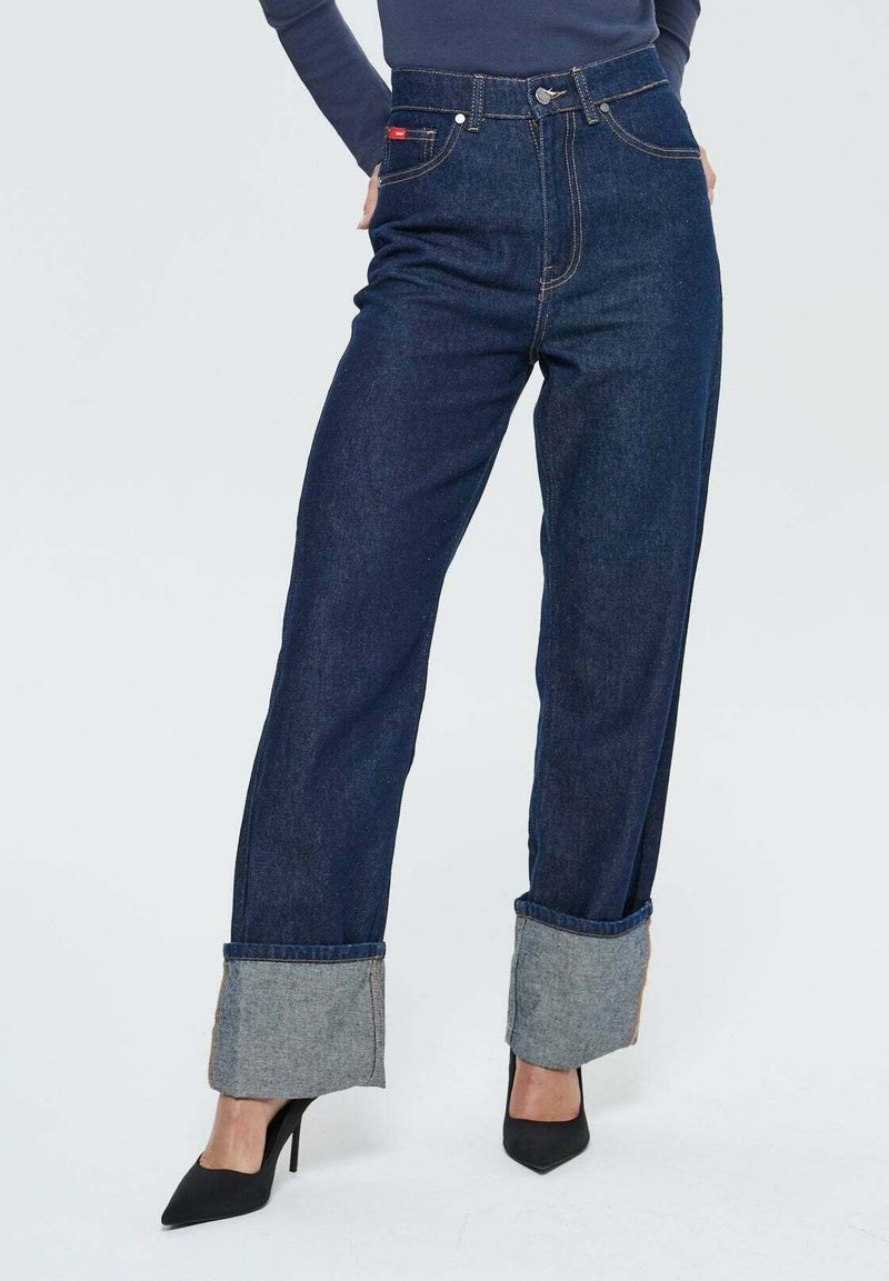 Femme portant un jean large à jambes larges bleu marine avec des talons hauts noirs et un haut à manches longues bleu marine devant un fond uni.