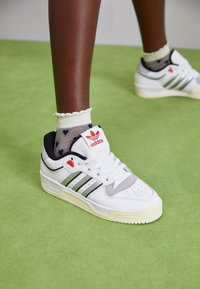 White leather sneakers with grey accents and green stripes, featuring a black collar and red logo on the tongue. Rubber sole.