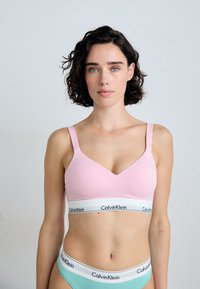 Světle růžový bezdrátový bralet s V-výstřihem, širokými ramínky a bílým pasem s logem "Calvin Klein". Hladký, elastický materiál.