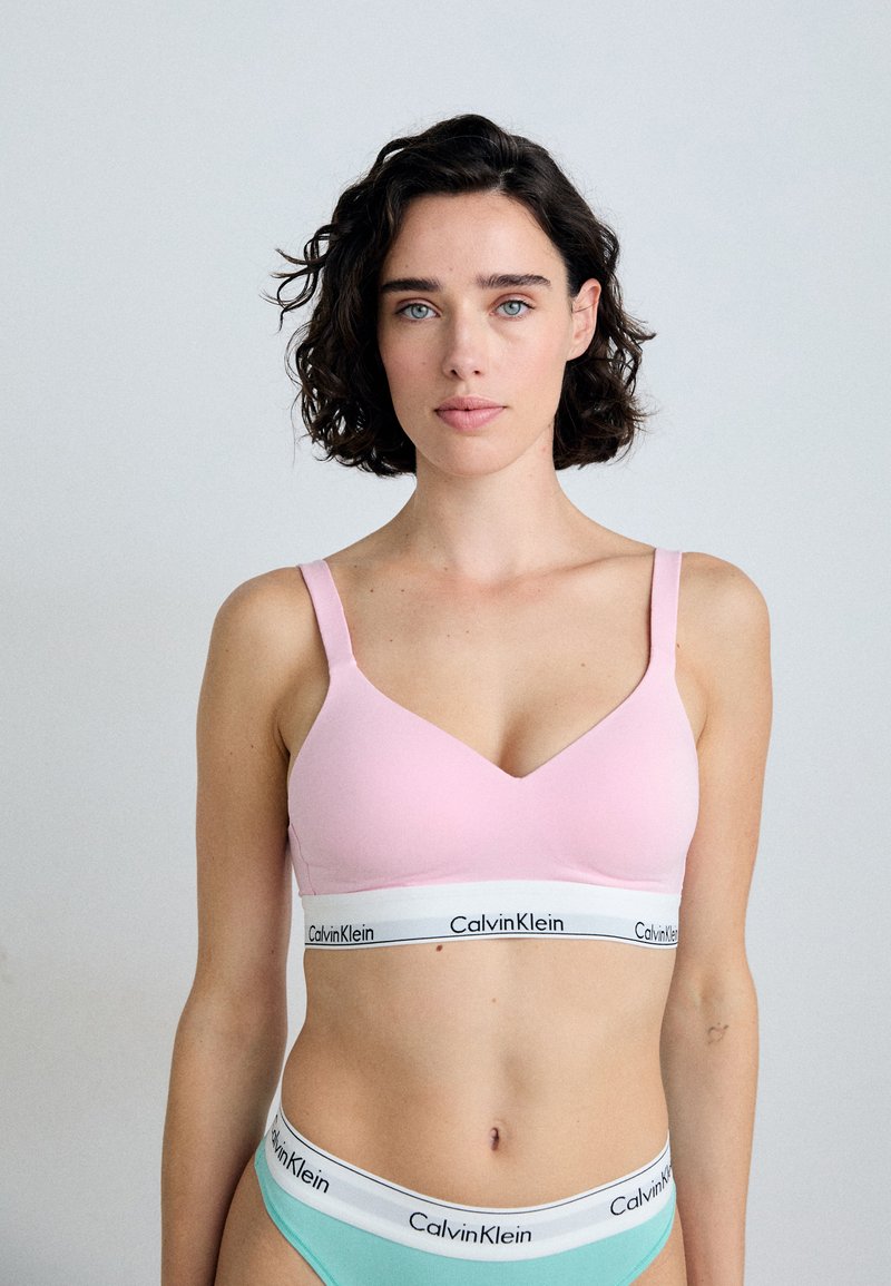 Světle růžový bezdrátový bralet s V-výstřihem, širokými ramínky a bílým pasem s logem "Calvin Klein". Hladký, elastický materiál.