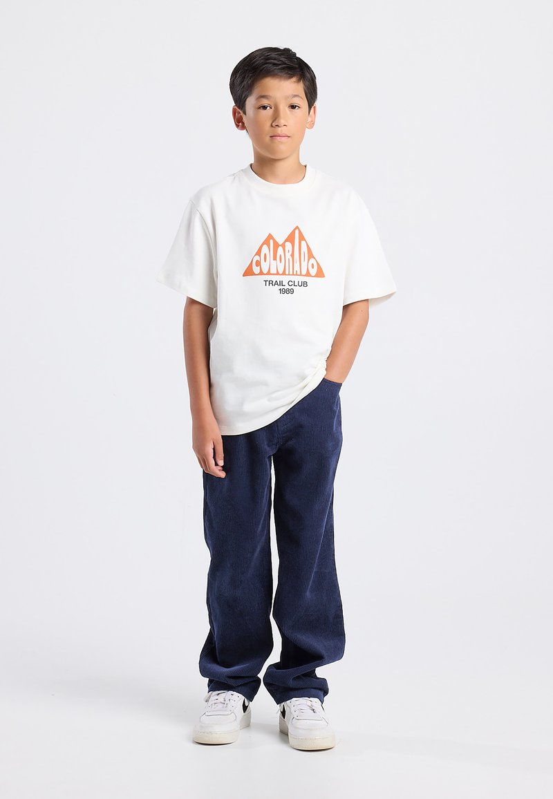 Jongen die staat met één hand in zijn zak, draagt een wit t-shirt met "Colorado Trail Club", marineblauwe broek en witte sneakers op een witte achtergrond.