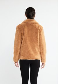 Brun, fuzzy fleecejakke med krave, lange ærmer og en afslappet pasform. Stoffet har et blødt, tekstureret udseende.