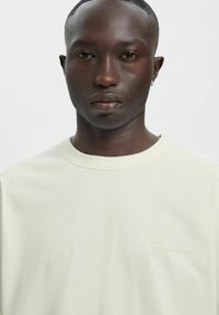 Flødefarvet bomulds t-shirt med rund halsudskæring, minimalistisk design med diskret trykt tekst på venstre side. Blød tekstur.