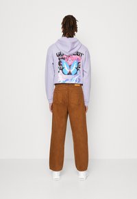 Lawendowa bluza z kapturem z kolorową grafiką motyla i napisem na plecach, zestawiona z brązowymi spodniami ze sztruksu i białymi trampkami.