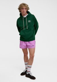 Green hoodie with white drawstrings and front pocket, paired with pink shorts. Model wears patterned socks and dark sneakers.