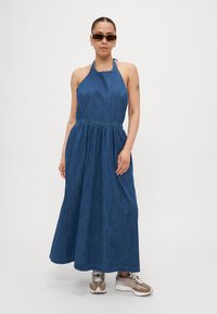 Denim halterkjole med tettsittende overdel og samlet midje, som går ut til en full skjørt. Farge: mørkeblå. Sammen med beige og grå sneakers.