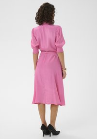 Robe rose avec manches bouffantes, ceinture à la taille et ourlet arrivant au genou. Texture de tissu lisse et silhouette ajustée, associée à des chaussures à talons hauts noires.