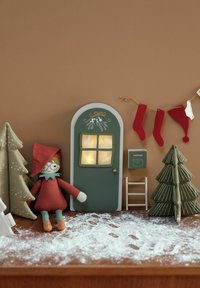 Poupée avec un chapeau rouge et un col bleu à côté d'une porte verte comportant une fenêtre. Autour, des arbres décoratifs, des bas de Noël et une poudre ressemblant à de la neige.