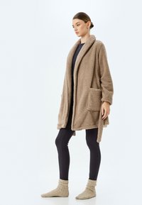 Maglione oversize, morbidamente voluminoso e color sabbia, con apertura frontale, colletto a scialle e due tasche anteriori, indossato sopra un top scuro e leggings, completato da calzini confortevoli.