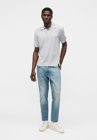 Άνδρας που στέκεται φορώντας ένα ανοιχτό γκρι polo με κοντά μανίκια, ανοιχτό μπλε τζιν, λευκές κάλτσες και λευκά αθλητικά παπούτσια σε απλό φόντο.